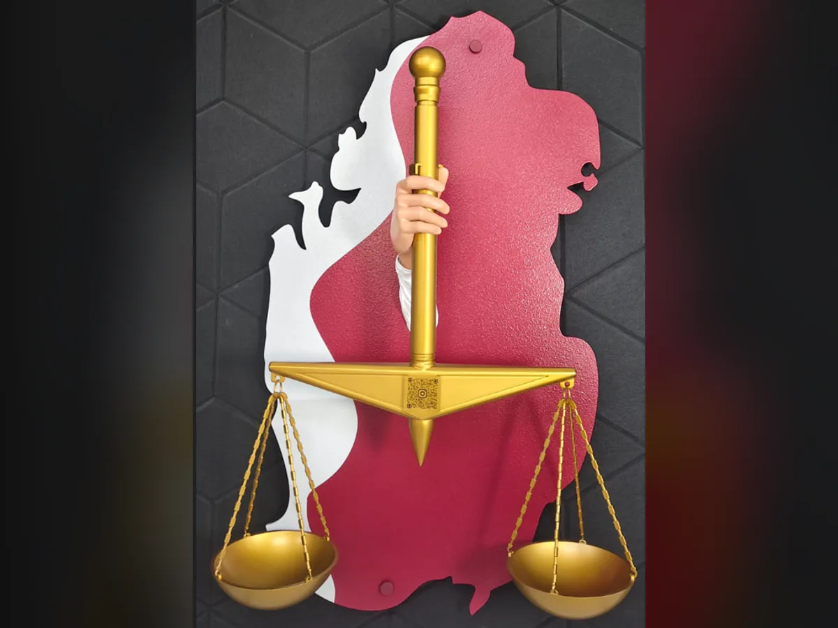 The Hand of Justice as delivered: a hand holding a golden scale out of a maroon and white map of Qatar, on a dark wall.
